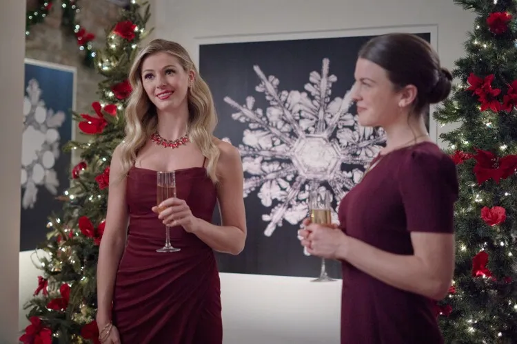 
This image features two women at a festive gathering, dressed in elegant burgundy dresses and holding champagne glasses. The woman on the left, adorned with a vibrant red necklace, is smiling and looking off to the side, likely engaging with someone outside the frame. Her companion appears more reserved, gazing thoughtfully at her glass. They stand in front of a striking black-and-white artwork of a snowflake, adding a sophisticated touch to the holiday-themed decor that includes a Christmas tree adorned with lights and red ornaments. The setting suggests a warm, celebratory atmosphere typical of holiday parties.