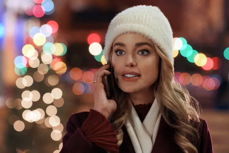 Hallmark Movie's "Operation Nutcracker": In this scene, we have a woman wearing a cozy white beanie and a burgundy coat, engaging in a phone conversation against a backdrop of festive lights that give off a merry, blurred sparkle. Her expression is attentive and somewhat curious, perhaps hinting at an intriguing or unexpected conversation as she navigates the holiday season's hustle and bustle. This type of image typically suggests a warm, festive storyline, possibly from a feel-good holiday film or advertisement.
