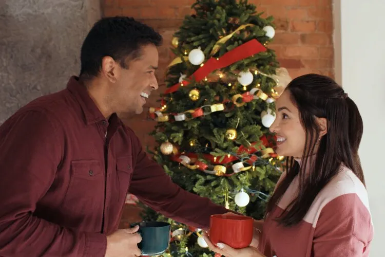 Hallmark Movie's "Operation Nutcracker": In this cozy holiday scene, a man and a woman share a joyful moment over cups of coffee by a beautifully decorated Christmas tree. The man is dressed in a burgundy shirt, and the woman in a soft pink sweater, both radiating warmth and happiness. Their expressions of laughter and engagement, along with the intimate setting and the festive backdrop, suggest a moment of lighthearted conversation and connection during the holiday season. This image perfectly captures the spirit of casual, heartfelt gatherings that are a hallmark of the festive period.
