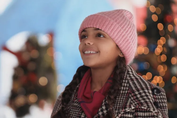 In this heartwarming scene, a young girl, adorned in a pink beanie and a cozy patterned coat, looks up with a look of wonder and joy. Her eyes are bright and her smile reflects genuine excitement, perhaps at the sight of something magical in the festive environment around her. The background, softly blurred with twinkling lights and holiday decorations, enhances the enchanting and joyful atmosphere typical of holiday celebrations. This image beautifully captures the innocence and happiness of childhood during the magical Christmas season.