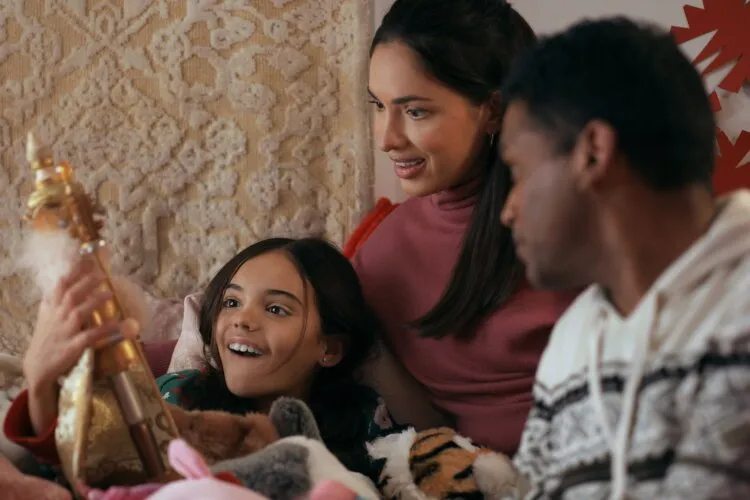 In this cozy scene, a family shares a joyful moment together during the holiday season. A young girl, surrounded by plush toys, holds up a nutcracker, her face lit up with delight. Both parents are focused on her, their expressions filled with affection and happiness. The mother, in a soft pink sweater, leans in closer with a smile, while the father, in a warm knit sweater, watches the interaction with a gentle gaze. The setting, rich with warm textures and a homely atmosphere, perfectly captures the essence of family gatherings and the special, simple moments of connection that define the holiday spirit.