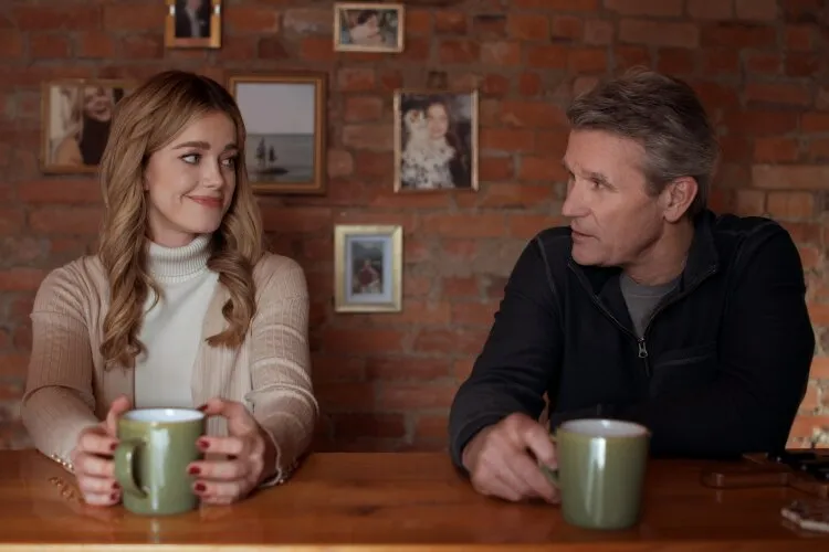 In this image, a man and a woman share a meaningful conversation over coffee in a warmly lit, rustic setting. The background, adorned with family photographs on a brick wall, adds a personal and intimate touch to the scene. The woman, dressed in a soft turtleneck sweater, gives a knowing smile, suggesting a shared joke or fond memory, while the man, dressed in a casual jacket, looks at her with interest and affection. Their relaxed postures and the cozy ambiance invite viewers into a moment of connection and understanding, common in heartwarming narratives where familial or close relationships are explored.