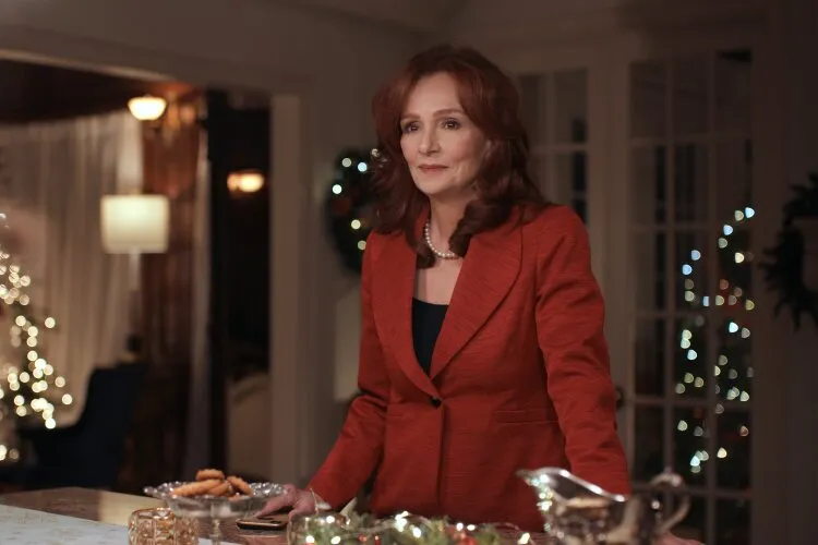 Hallmark Movie's "Operation Nutcracker": In this image, a woman stands poised at a festively decorated table in a warmly lit room, evoking a sense of holiday elegance. Dressed in a stylish red blazer, she exudes a calm and composed demeanor, which may indicate her role as a host or an important figure in a holiday gathering. The background, softly illuminated with Christmas lights and tasteful decorations, adds to the festive atmosphere. Her expression, attentive and slightly introspective, suggests she is preparing for an event or reflecting on a moment important to the narrative, potentially involving family traditions or holiday celebrations.