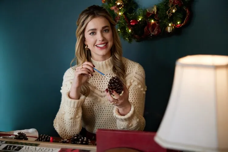 Hallmark Movie's "Operation Nutcracker": In this festive scene, a woman engages in holiday crafting, painting a pine cone with a cheerful smile. Dressed in a cozy, knitted sweater, her joyful expression and the creative setting suggest she's deeply involved in making holiday decorations. The background, adorned with a colorful garland and set against a rich teal wall, provides a warm and inviting atmosphere. This image captures a moment of holiday preparation, likely part of a storyline that emphasizes traditions, creativity, and the personal touch of handmade decor in celebrating the season.