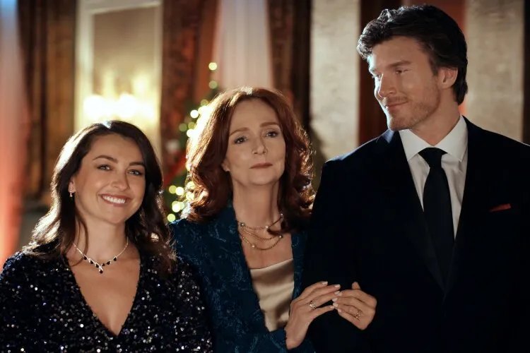 In this elegant holiday scene, three individuals—two women and a man—pose together in formal attire, exuding a sense of familial connection or close friendship. The woman in the center, dressed in a sophisticated blue gown, stands with a confident and composed expression between a younger woman in a sparkling navy dress and a man in a sleek black suit. Their expressions, ranging from gentle smiles to fond gazes, suggest a moment of celebration or a significant event, possibly at a formal holiday gathering. The backdrop, illuminated by soft, warm lights and holiday decor, enhances the festive and refined atmosphere of the scene.