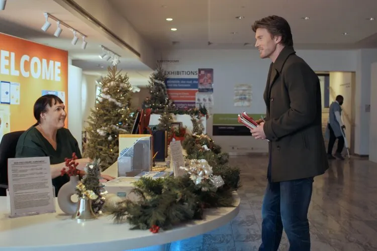 Hallmark Movie's "Operation Nutcracker": In this scene, a man stands at a reception area, engaging with a receptionist during the holiday season. The man, dressed in a stylish coat and jeans, holds books, suggesting he might be visiting for an event or meeting. The receptionist, seated behind a festively decorated desk with Christmas trees and garlands, greets him with a welcoming smile. This setting, likely in a community center or similar venue, is adorned with holiday decorations, adding a festive touch to what appears to be a pleasant and friendly interaction. This image captures a moment of everyday warmth and seasonal hospitality.