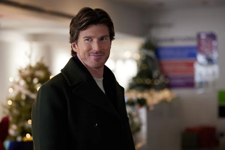In this image, a man offers a warm smile in a festive indoor setting, signaling a moment of pleasant interaction or arrival at a holiday event. Dressed in a stylish green coat over a casual sweater, his relaxed yet sophisticated attire suits the festive backdrop, which includes holiday decorations and trees. His confident and friendly demeanor suggests he's either a key figure in the narrative or enjoying a casual visit to a community space adorned for the holiday season. The setting, with its welcoming atmosphere and seasonal decor, emphasizes a theme of community and festive gatherings.