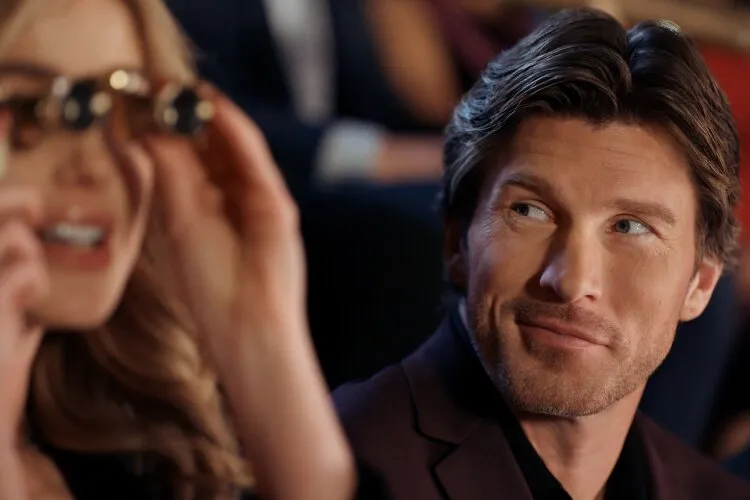 Hallmark Movie's "Operation Nutcracker": This photo captures a charming moment between a man and a woman at a social event. The man, wearing a dark burgundy suit, looks at the woman with a smile and an expression of admiration or amusement. Meanwhile, the woman, slightly out of focus in the foreground, is adjusting her sunglasses with a playful gesture. The blurred background suggests they are in a bustling, elegant setting, possibly during a festive gathering or a special event, adding a layer of sophistication and warmth to the scene.