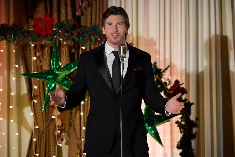 In this festive scene, a man dressed in a sharp black suit stands confidently at a microphone, addressing an audience at a holiday event. His poised demeanor and engaging smile suggest he is either making a speech or serving as an emcee. The background is festively decorated with holiday lights, poinsettias, and large green stars, creating a vibrant and celebratory environment. This image captures a moment of leadership and charisma, highlighting his role in facilitating the festivities and enhancing the holiday spirit among the attendees.