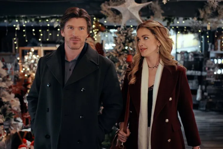 Hallmark Movie's "Operation Nutcracker": In this festive image, a man and a woman are strolling through a holiday market, surrounded by Christmas decorations and twinkling lights. The man is dressed in a classic dark peacoat, while the woman wears a stylish burgundy coat with a white scarf. Both are looking off to the side, perhaps captivated by something in the market, adding a sense of wonder and holiday excitement to the scene. The beautifully decorated background with sparkling lights and Christmas ornaments sets the perfect winter holiday atmosphere.