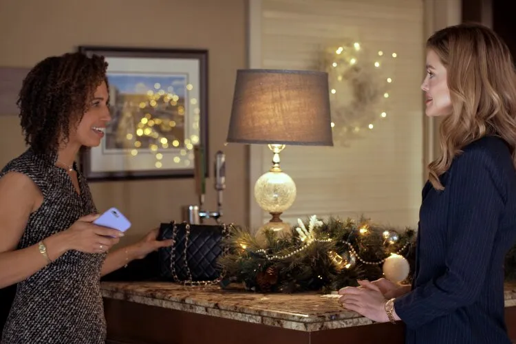 This scene captures a warm and festive interaction between two women in a beautifully decorated room for the holiday season. The woman on the left, holding a smartphone, seems to be sharing something interesting or humorous on her screen with the woman on the right, who listens attentively. The room is adorned with a Christmas garland and lights on the console behind them, which enhances the cozy, festive atmosphere. This setting likely suggests a moment of friendship and joyful sharing during the holiday celebrations.