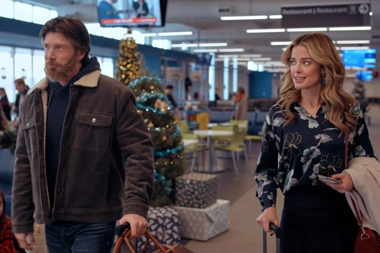 Hallmark Movie's "Operation Nutcracker": In this image, a man and a woman are captured walking through an airport during the holiday season. The man, dressed in a rugged winter jacket, carries a brown leather bag, while the woman, sporting a chic floral blouse, pulls along a suitcase. The background features festive Christmas decorations, including trees and wrapped gifts, creating a cheerful and bustling travel scene. Their expressions suggest they are in the midst of a conversation, possibly about their travel plans or the holiday season, adding a layer of engagement to the scene. This setting likely emphasizes themes of journey and reunion, common in festive narratives.