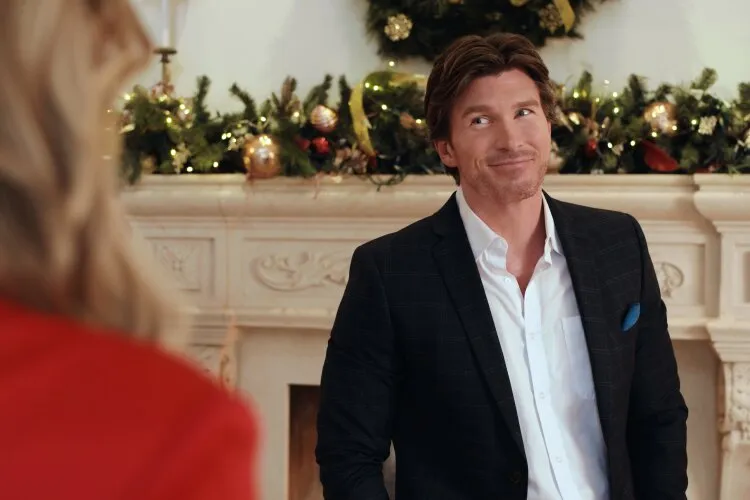 This image features a man dressed in a stylish blazer and shirt, giving a charming smile to someone just out of frame, likely during a festive conversation. Behind him, a beautifully decorated Christmas mantel, adorned with glowing lights and richly colored ornaments, sets a holiday-perfect scene. His expression suggests a light-hearted, perhaps playful moment, common in feel-good holiday movies where romance or family joy is a central theme. This setting evokes the warmth and coziness of Christmas, ideal for scenes of holiday gatherings and heartfelt exchanges.