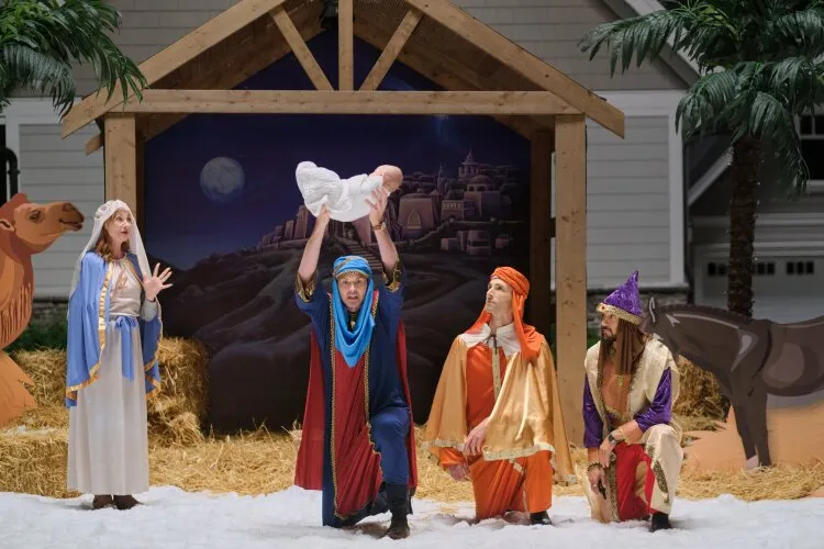 Four people are dressed in nativity costumes, performing a scene in front of a stable backdrop. One man is lifting a baby wrapped in white cloth high in the air, while the other three characters look on with expressions of awe and joy. The scene is surrounded by hay, palm trees, and painted cutouts of animals, including a camel and a donkey.