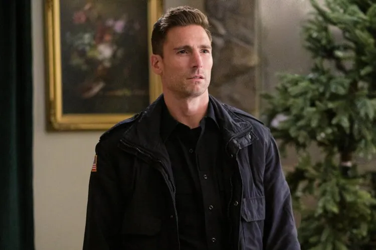 From Hallmark's "Three Wise Men and a Baby": A man is standing indoors, wearing a dark jacket and shirt. He has a serious expression on his face, suggesting he is deep in thought or concerned about something. In the background, there is a framed painting and some greenery, creating a homey atmosphere. The man's posture and expression convey a sense of determination or resolve.