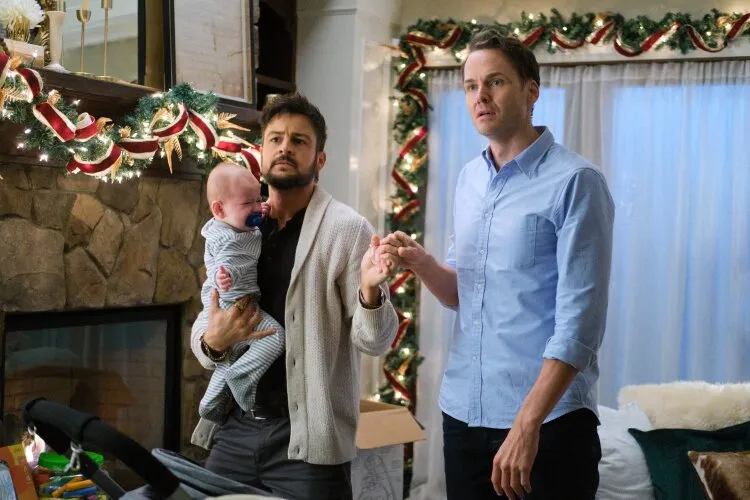 Two men are standing in a festively decorated living room, one holding a crying baby. The man holding the baby looks concerned while the other man has a similarly worried expression. The room is adorned with Christmas garlands and lights, and a stone fireplace is visible in the background. The scene captures a moment of distress during an otherwise festive setting.