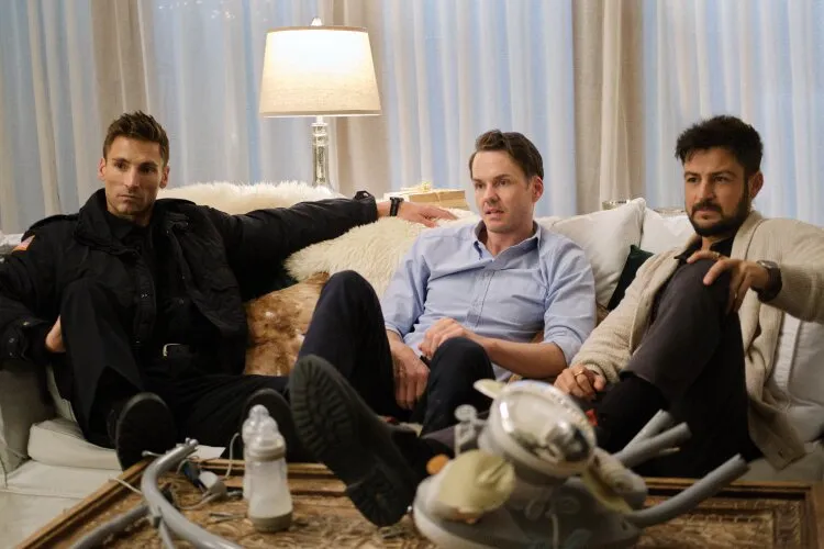 From Hallmark's "Three Wise Men and a Baby": Three men are sitting on a couch in a cozy living room. One man, dressed in a dark jacket, is lounging with his arm on the back of the couch. The other two men, one in a light blue shirt and the other in a beige sweater, are sitting more upright, looking engaged or concerned. The coffee table in front of them is cluttered with baby items, including a bottle and parts of baby furniture. A lamp provides warm lighting in the background, adding to the comfortable atmosphere.