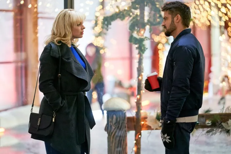 A woman with blonde hair, wearing a dark coat, is having a conversation with a man holding a red coffee cup. They are standing outdoors in a festive setting with Christmas lights and decorations. The atmosphere is warm and cheerful, with both characters engaged in what appears to be a meaningful conversation. The background is softly lit with holiday decor, adding to the seasonal charm.
