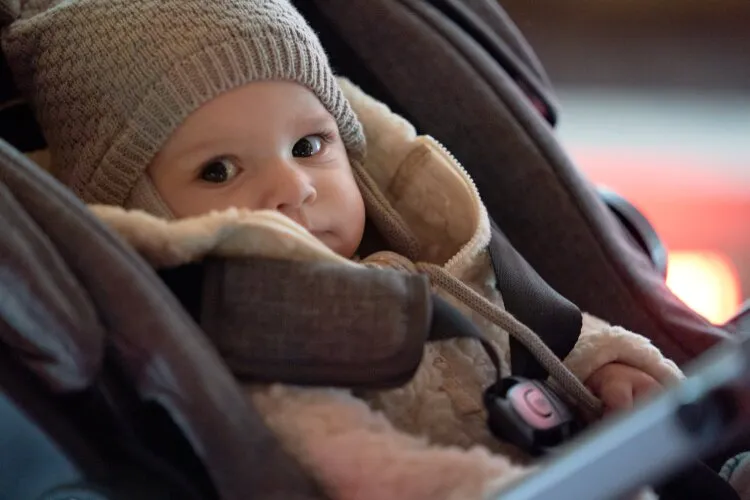 From Hallmark's "Three Wise Men and a Baby": A baby is bundled up in a cozy jacket and knitted hat, sitting in a stroller. The baby has a curious and wide-eyed expression, looking directly at the camera. The setting appears to be indoors, with soft lighting that highlights the baby's adorable features and warm outfit.