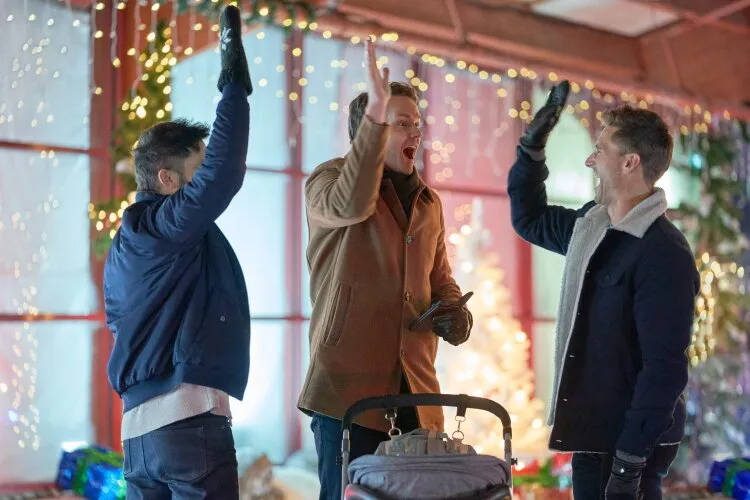 Three men are standing indoors, giving each other high-fives with excitement. They are dressed in winter clothing, and in the foreground, there is a baby stroller. The background features festive decorations, including string lights and a decorated Christmas tree.