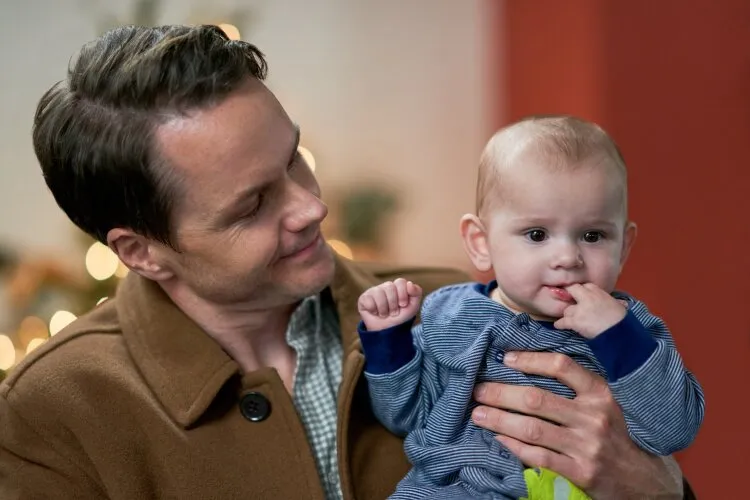 A man is holding a baby dressed in a striped outfit. The man, wearing a brown coat, is smiling warmly at the baby, who has a curious expression and is touching its own mouth. The background is softly lit, with festive decorations adding to the cozy and heartwarming atmosphere.