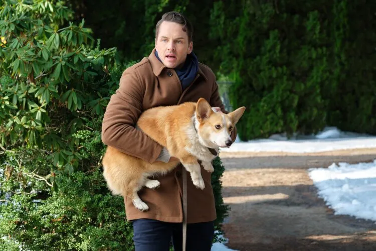 From Hallmark's "Three Wise Men and a Baby": A man wearing a brown coat is outdoors, holding a Corgi dog in his arms. He has a concerned expression on his face. The background features greenery and a snowy path, suggesting a winter setting.