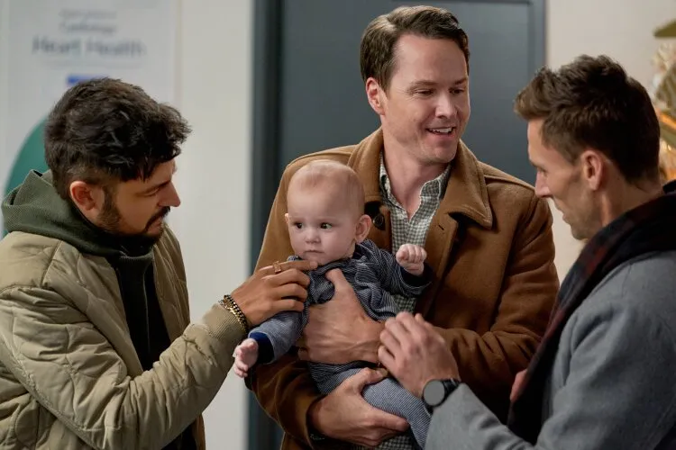 Three men are gathered around a baby, with one man holding the baby while the other two gently interact with it. They are indoors, dressed in casual winter clothing. The scene appears warm and caring, with the men smiling and focused on the baby. In the background, there is a poster and part of a Christmas tree.