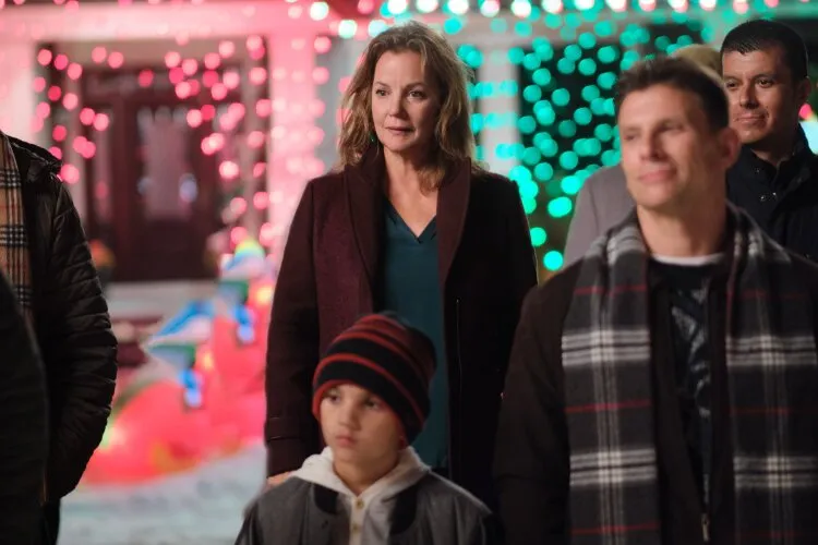 A group of people, including a woman in a dark coat, a man with a plaid scarf, and a child wearing a striped beanie, are standing outdoors at night. They are surrounded by colorful Christmas lights and decorations, creating a festive atmosphere. The expressions on their faces suggest they are watching something intently.