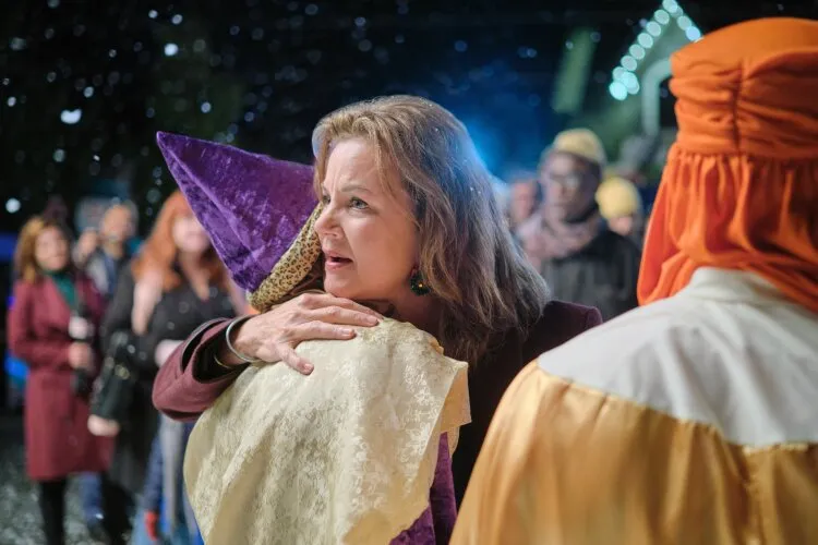From Hallmark's "Three Wise Men and a Baby": A woman is hugging a person dressed in a purple hat and a cream-colored robe. They are outdoors at night, surrounded by people and festive decorations. The scene is set during winter, as snow is falling, adding to the festive atmosphere. The woman looks emotional and comforting, with other people blurred in the background.