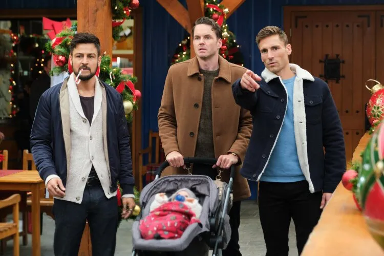 From Hallmark's "Three Wise Men and a Baby": Three men are standing together, with one pushing a baby stroller covered with a red blanket. They are dressed in casual winter clothing and are in a festive setting with Christmas decorations around them. One man is pointing at something off-camera with a serious expression, while another has a candy cane in his mouth, and the third looks concerned. They are in what appears to be a cozy, decorated outdoor market or gathering area.