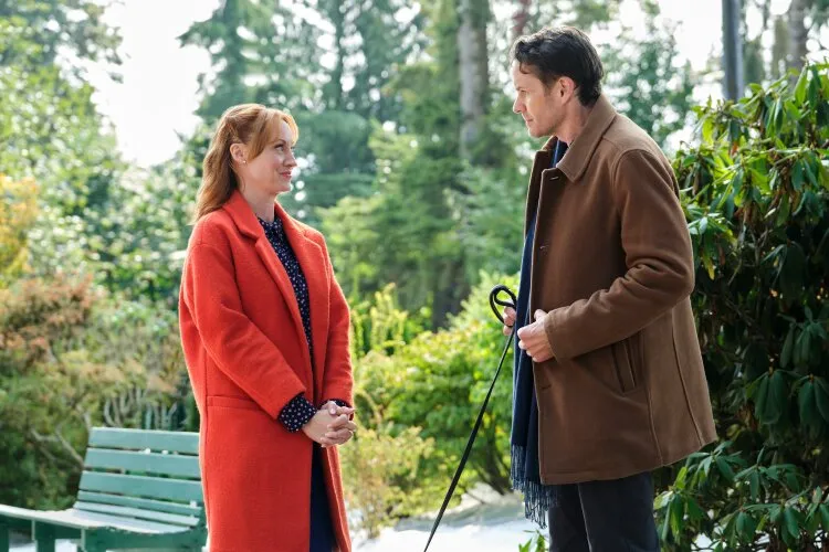 A man and a woman are standing outdoors, facing each other and engaging in conversation. The woman is wearing a bright red coat, while the man is dressed in a brown coat and holding a leash. The background features lush greenery, suggesting a park or garden setting, with a green bench visible to the side. Both appear to be in a relaxed and pleasant mood.