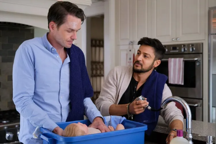 Two men are in a kitchen, washing a baby in a large blue plastic tub placed in the sink. One man, with a towel draped around his neck and a concerned expression, is gently holding the baby. The other man is using a sprayer faucet, looking at his friend with a reassuring smile. The kitchen setting is modern, with white cabinets and stainless steel appliances in the background. The scene is intimate and caring, depicting a moment of shared responsibility and friendship.