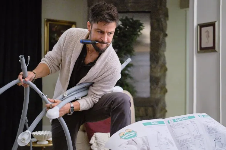 A man is attempting to assemble a piece of baby furniture, holding several parts and a tool in his mouth. He looks focused and slightly frustrated, with an instruction manual spread out in front of him. The setting is a cozy living room, with a glimpse of a stone fireplace and some artwork on the walls. The scene humorously captures the challenges of putting together baby gear.