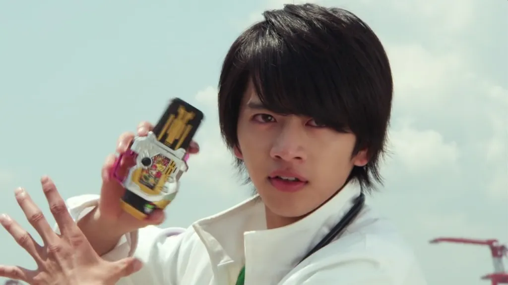 Emu Hojo from Kamen Rider Ex-Aid, dressed in a white lab coat, holds up a Gamer Driver with determination in his eyes. His dark hair and serious expression highlight his resolve to transform and protect. The background features a clear sky, adding intensity to the moment.