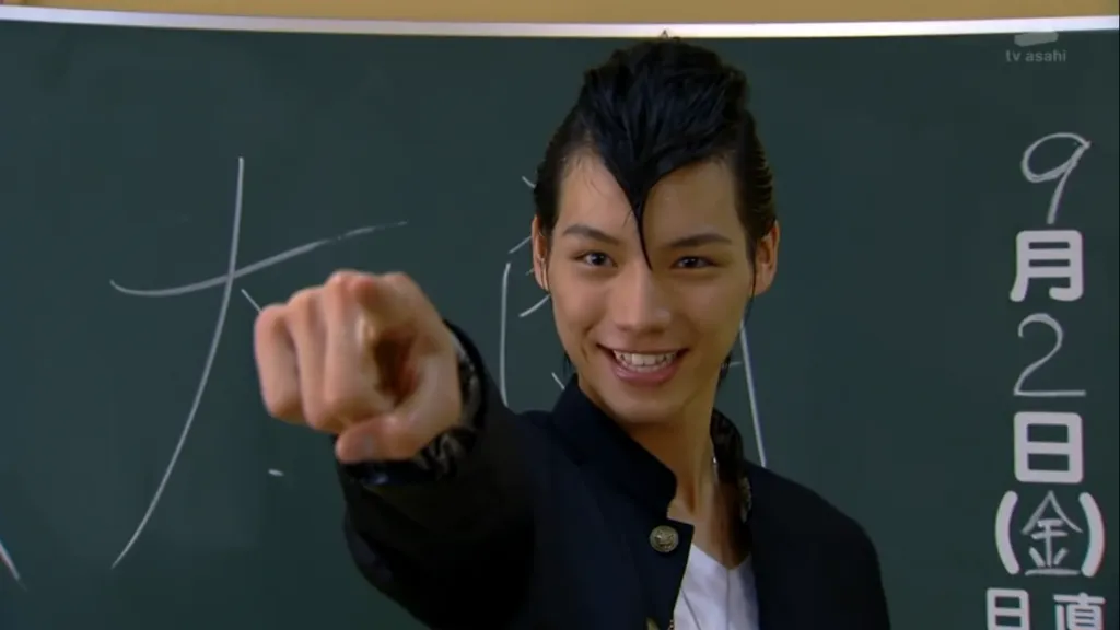 Gentaro Kisaragi from Kamen Rider Fourze stands confidently in front of a classroom blackboard, pointing directly ahead with a determined and friendly smile. His slicked-back hair and school uniform reflect his unique and charismatic personality. The scene captures Gentaro's enthusiasm and commitment to forming bonds of friendship.