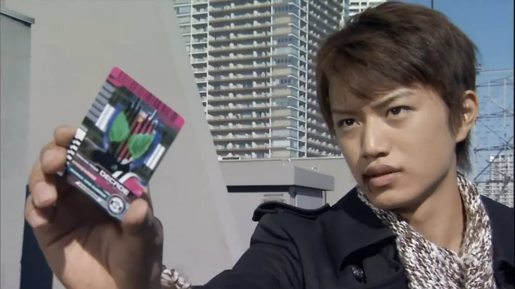 Tsukasa Kadoya, the protagonist of Kamen Rider Decade, holds up a transformation card with a serious expression. The urban backdrop emphasizes his readiness for action as he prepares for another henshin.