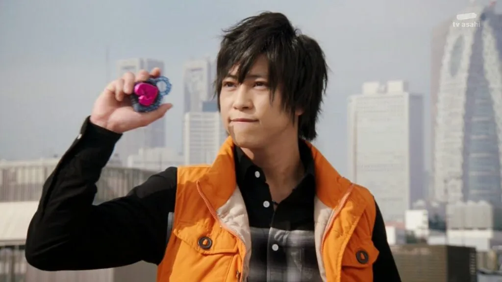 Kota Kazuraba from Kamen Rider Gaim stands determinedly on a rooftop, holding up a pink Lockseed. Dressed in his signature orange vest over a black shirt, Kota looks ready to transform, with the cityscape of tall buildings in the background reflecting his commitment to protect his friends and city.