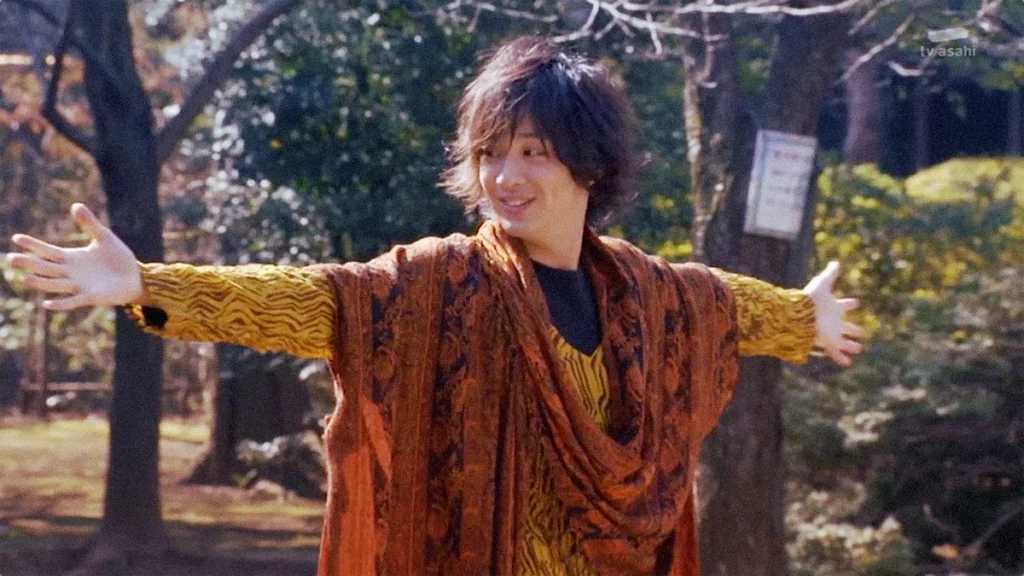 Eiji Hino from Kamen Rider OOO stands with his arms wide open, smiling warmly. He's dressed in his signature layered, colorful attire, set against a backdrop of lush greenery in a park. The scene exudes a sense of freedom and optimism, capturing Eiji's adventurous and kind-hearted nature.