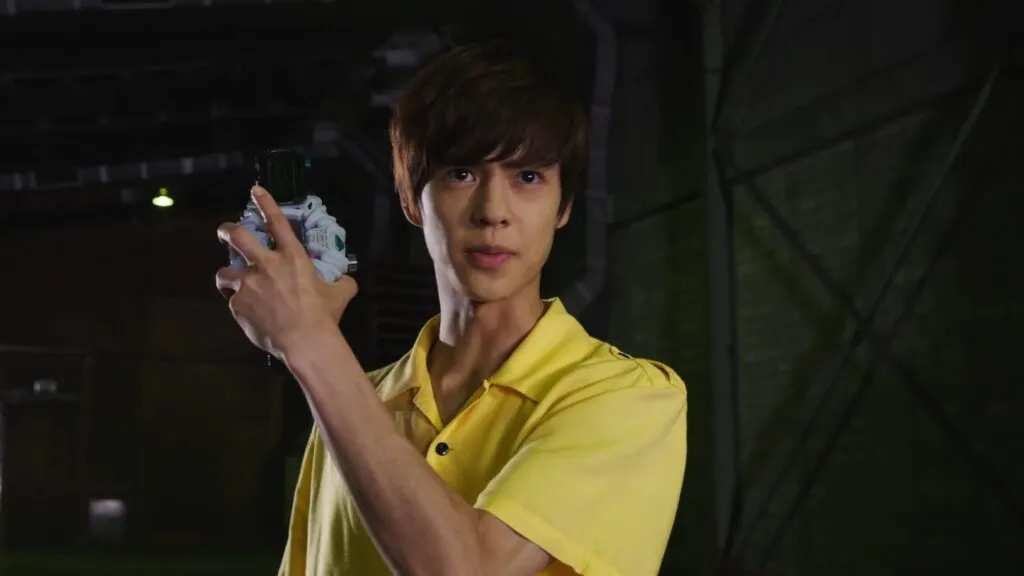 Sougo Tokiwa from Kamen Rider Zi-O, dressed in a yellow shirt, holds up a Ridewatch with a determined expression. The background is dark and industrial, highlighting the tension and importance of the moment.