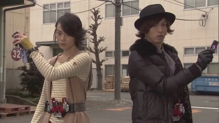 Shotaro Hidari and Phillip, the protagonists of Kamen Rider W, stand ready for action. Both are holding their transformation devices, prepared for their iconic double transformation. The urban setting adds to the atmosphere of their detective-themed adventures.