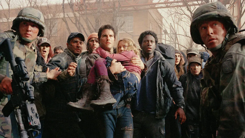 A dramatic scene from the movie 'War of the Worlds' featuring Tom Cruise as the character Ray Ferrier. Ray is seen carrying his young daughter, Rachel, portrayed by Dakota Fanning, while surrounded by a crowd of distressed people and soldiers. The setting appears chaotic, with a sense of urgency and tension. Soldiers in camouflage uniforms and helmets are on either side, and the background shows an urban environment with bare trees and buildings. One of the iconic New Jersey Film Locations.