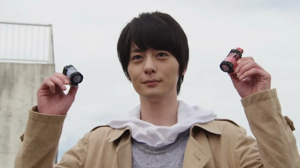 Kiryu Sento from Kamen Rider Build stands confidently, holding up two Full Bottles, ready for transformation. Dressed in a tan coat over a grey sweater with a white hoodie underneath, Sento's determined expression shows his readiness to protect and fight for justice. The background features a simple outdoor setting with a cloudy sky.