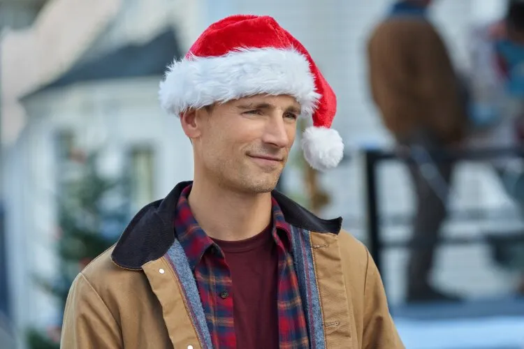 Andrew Walker is wearing a Santa hat and a casual winter outfit, including a brown jacket over a plaid shirt. He is outdoors, with a blurred background suggesting a festive setting. This image is from the Hallmark movie "Christmas Island."