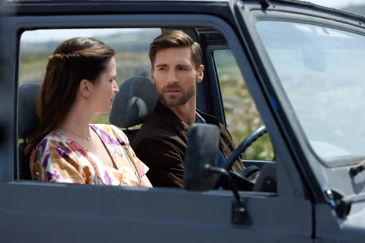 A scene from Andrew Walker's For Love & Honey on Hallmark. A man and woman are sitting in a car, facing each other and engaged in a conversation. The man, with short brown hair and a beard, is in the driver's seat wearing a dark jacket. The woman, with long brown hair pulled back, is in the passenger seat wearing a colorful floral blouse. They appear to be sharing an intimate moment, with the scenery outside the car suggesting a picturesque, rural setting.