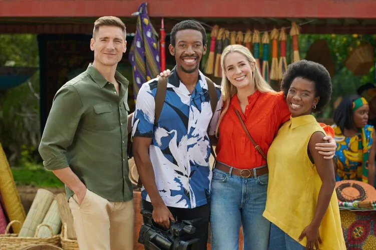 Andrew Walker is seen with three other people, all smiling and standing close together, suggesting camaraderie. The group is outdoors in a colorful setting with market stalls and vibrant fabrics in the background. This image is from the Hallmark movie "Safari Romance."