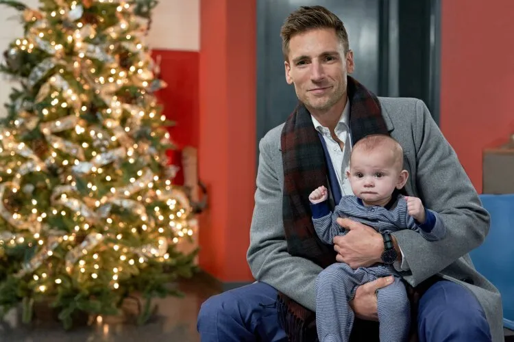 Andrew Walker is seen sitting and holding a baby in his arms, with a warmly lit Christmas tree in the background. The setting exudes a festive holiday atmosphere, and both are dressed in cozy, winter-appropriate clothing. This scene is from the Hallmark movie "Three Wisemen and a Baby."