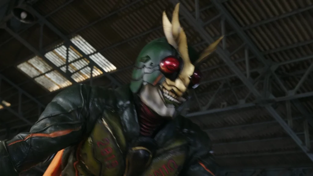 A scene from the Tokusatsu series featuring Kamen Rider Another Agito. The character is in full costume, which includes a dark green and black armor with a rugged, organic design. The helmet features large, red compound eyes and two prominent golden horn-like structures, resembling insect antennae. The character has a fearsome and menacing appearance, with a skeletal, almost monstrous mouthpiece that adds to the intimidating look. The chest armor is detailed and worn, indicating intense battles. The background is an industrial setting with metal beams and a ceiling, suggesting a warehouse or factory environment. The overall atmosphere is intense and foreboding, showcasing the unique and powerful design of Kamen Rider Another Agito.