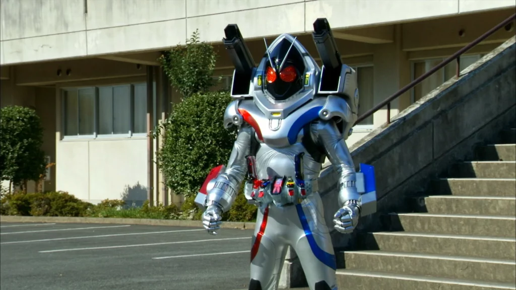 A scene from the Tokusatsu series featuring Kamen Rider Fourze in his Magnet States form. The character is in full costume, characterized by a futuristic, metallic armor with silver, blue, and red accents. The helmet has large, red compound eyes and a sleek, angular design with a black visor. Mounted on his shoulders are large, cannon-like structures, enhancing the high-tech appearance. The chest and arms have a robust, armored look, and a belt around his waist holds various gadgets and devices. The character stands in front of a building with stairs and greenery, creating a contrast between the modern, mechanical suit and the everyday environment. The overall impression is one of technological advancement and readiness for action, fitting the heroic and powerful nature of Kamen Rider Fourze.