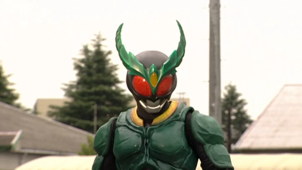 A scene from the Tokusatsu series featuring Kamen Rider Gills. The character is in full costume, characterized by a predominantly green armor with a sleek, organic design. The helmet features large, red compound eyes and a prominent green horn-like structure that resembles dragonfly wings, enhancing the insect-like motif. The chest armor has a textured, almost muscular appearance, adding to the character's dynamic and formidable look. The background is an outdoor setting with trees and buildings, creating a natural, yet urban environment. The overall atmosphere is intense and showcases the unique and powerful design of Kamen Rider Gills.