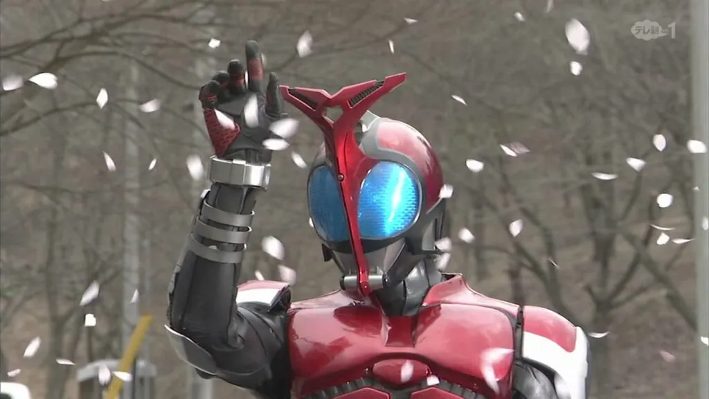 A scene from the Tokusatsu series featuring Kamen Rider Kabuto. The character is in full costume, which includes a metallic red and black armor with a beetle-like design. The helmet has large, glowing blue eyes and a distinctive red "horn" on top. The character is posed with one hand raised, fingers slightly curled, as cherry blossom petals fall around him. The background is an outdoor setting with bare trees, emphasizing a serene yet powerful atmosphere.
